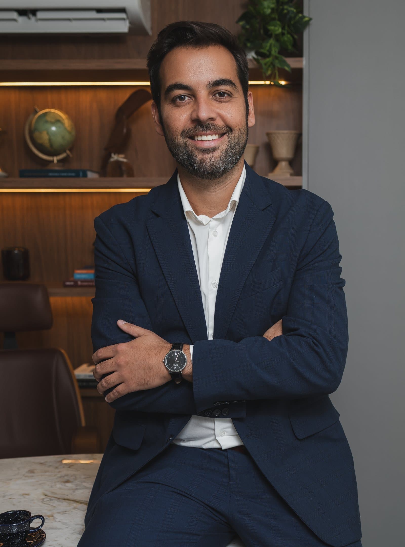 Dr. Gabriel Bastos sentado no consultório, de terno azul, com os braços cruzados.